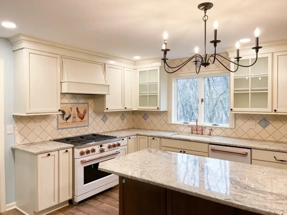 A warm, traditional kitchen featuring cream-colored cabinetry and a custom backsplash with a diamond tile pattern accented by decorative blue tiles. The centerpiece above the stove includes a charming rooster tile mural, adding a touch of farmhouse character to the space. Granite countertops with light veining complement the soft tones of the cabinetry, while a large island provides ample workspace. A classic black wrought-iron chandelier hangs over the kitchen, adding elegance and contrast to the bright, inviting design. Large windows allow natural light to flood the space, highlighting the craftsmanship and details of the kitchen.