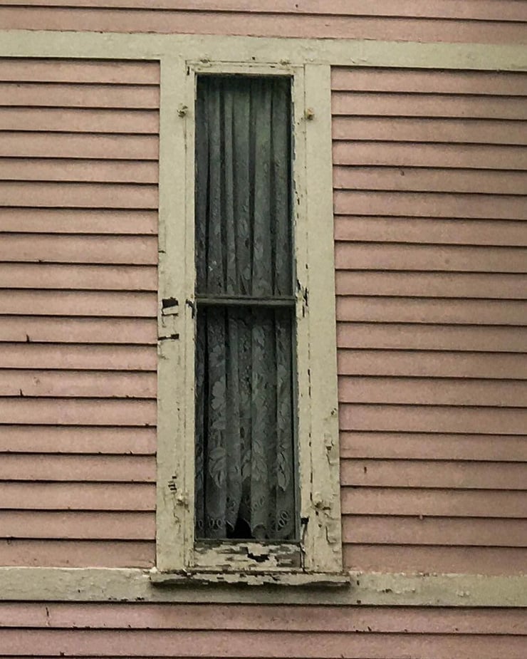 Detail view of an original single-hung wood window on a historic Fayetteville, NY home before replacement. Features chipped paint, worn trim, and lace curtain, captured prior to McClurg Remodeling’s custom window restoration.