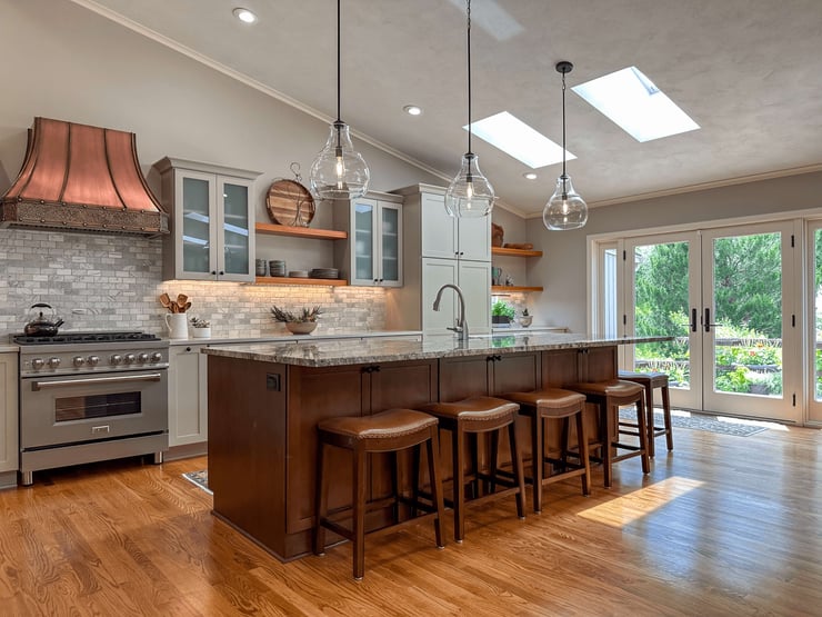 Remodeled kitchen with layered lighting design featuring glass pendant lights over island, recessed ceiling lights, under-cabinet lighting, and natural light from skylights and French doors overlooking lush garden patio.