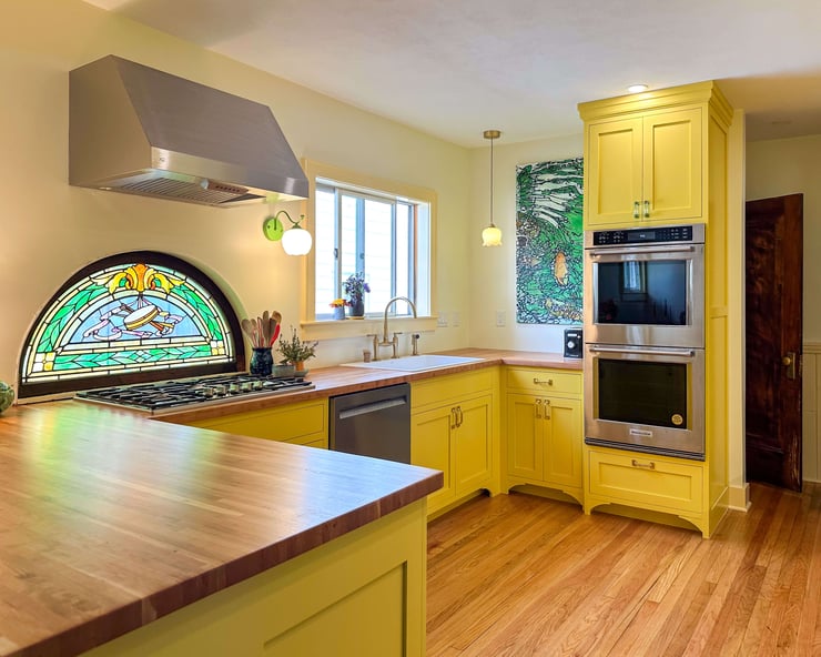 Charming kitchen with bright yellow cabinets, butcher block countertops, layered lighting including recessed, pendant, and wall-mounted sconces, and a decorative stained-glass window over the cooktop.