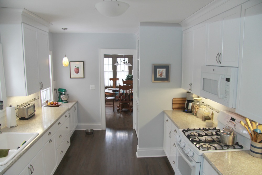 A galley kitchen was brought up-to-date and restored to reflect the original character of a home built in 1929 by adding white painted Shaker-style cabinets, white oak wood flooring, white appliances, a white cast-iron sink, laminate counters, tile backsplash and new lighting fixtures.
