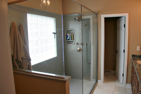 This walk-in shower without doors has a glass surround with a kneewall. The window is diamond cut glass set on a smooth plane within the shower so that water will not accumulate on the framing. A custom Onyx shower base with a low threshold and Onyx shower wall panels with a stone tile gloss finish (Slate Stone 12-by-12 inch tile design in “Spanish Moss”) were used for the shower. The shower includes a wall niche, a shower seat, a wall-mounted rainfall showerhead, a handheld showerhead and a sealed can light in the ceiling.