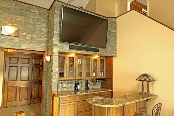 An oak bar with a stunning granite counter was built into the room. Glass door cabinets display the barware and a stemware rack holds wine glasses. A shelf was built under the top cabinets to accommodate a cable box. Cabinets are from Jim Bishop. The stain is a custom stain requested by the homeowners to match existing oak features. A flat screen television was mounted over the bar.